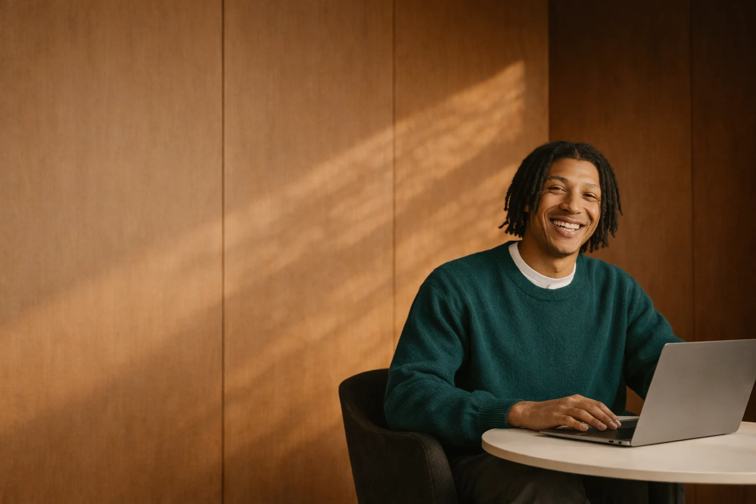 Smiling young man with dreadlocks wearing a green sweater sitting at a round table using a laptop.