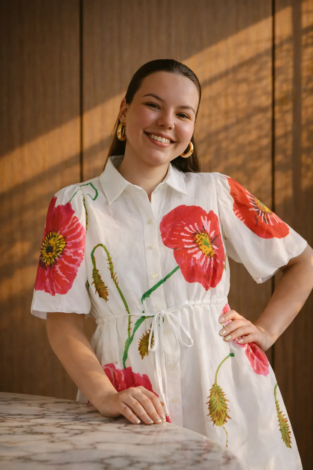 Smiling woman with long dark hair wearing gold hoop earrings and a white dress with large red floral prints, leaning on a marble countertop.